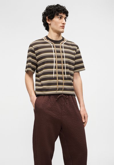 Άνδρας που φοράει καφέ και μπεζ κοντομάνικη ριγωτή μπλούζα με λευκά και μπεζ κορδόνια που κρέμονται, και υφασμάτινο σκούρο καφέ παντελόνι με υφή.