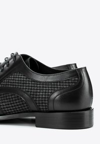 Zapatos de vestir negros con parte superior de tejido texturizado, acentos de cuero, punta redonda y suela plana de goma. Diseño con cordones y seis ojales visibles.