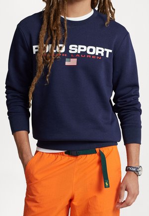 Granatowy sweter z napisem "POLO SPORT" w białym i czerwonym kolorze, z amerykańskim emblematem flagi; zestawiony z pomarańczowymi szortami i zielonym paskiem.