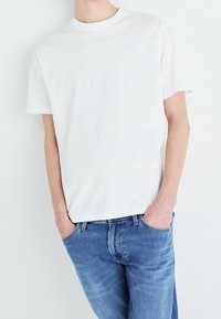 Vitt bomullströja med rund hals, korta ärmar och en diskret logotyp tryckt på bröstet, matchad med ljusblå denimjeans.