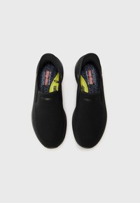 Black slip-on shoes with a textured mesh upper and a cushioned insole. The sole is sleek and round, featuring yellow accents inside.