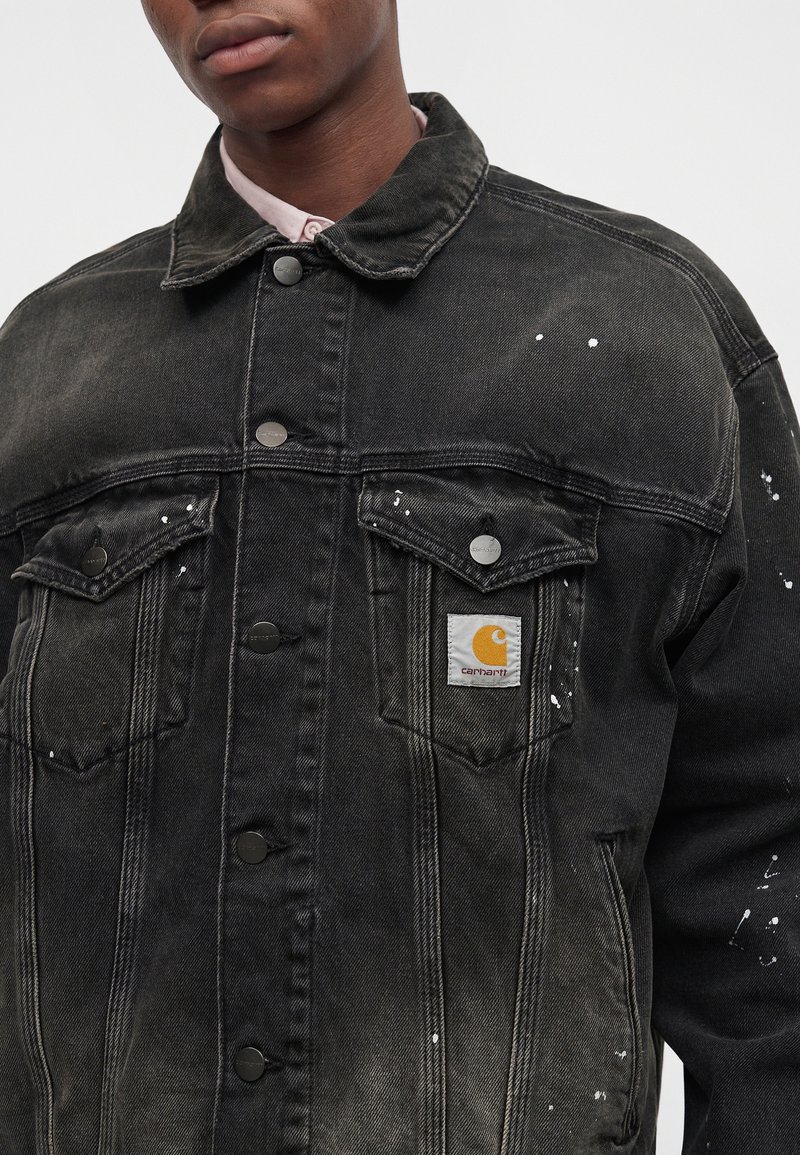 Young person wearing a black Carhartt denim jacket with white paint splatters and metal buttons, over a light pink collared shirt.