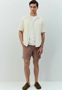 Korte mouwen roomkleurig shirt met een gestructureerd patroon, gecombineerd met bruin gestreepte shorts en olijfgroene instapschoenen.