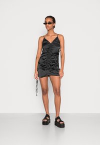 Vestido mini de satin preto com alças ajustáveis, detalhe franzido ao centro e um laço de cordão de um lado, combinado com sandálias pretas chunky.