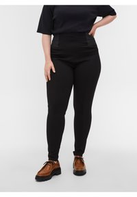 Pantalons noirs taille haute avec des panneaux en tissu contrasté à la taille, présentant une texture lisse et une coupe fuselée. Chaussures brunes avec une semelle épaisse.