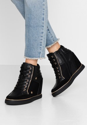 Person iført lyseblå korte jeans og sorte kilehælssneakers med guldøjer og sidelommer på en grå gulv.