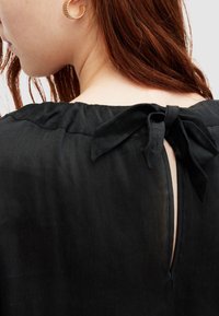 Gros plan sur l’épaule d’une femme aux cheveux roux portant un haut noir noué en nœud dans le dos et une boucle d’oreille en anneau doré.