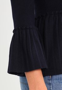 Mörkblå stickad topp med ribbad textur, som har vida ärmar med volangkanter och en utställd design vid nederkanten.