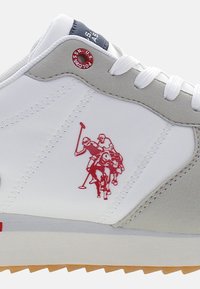Sneaker bianca con accenti grigi, caratterizzata da un ricamo di un giocatore di polo rosso. Suola testurizzata e punta arrotondata; lacci fissati su una linguetta imbottita.