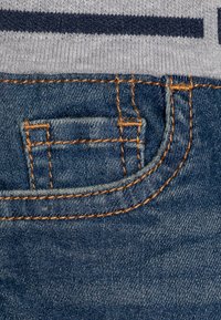 Denimstoff, dunkelblaue Farbe, mit einer kleinen Fronttasche und kontrastierenden orangen Nähten sowie einem grauen Bund mit marineblauen Streifen.