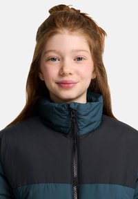 Polstret jakke med høy krage, med et fargeblokk-design i mørk teal og svart, og glidelås foran. Myk tekstur og isolerende materiale.