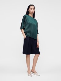 Femme portant un haut plissé vert foncé, un short noir ample longueur genou et des talons pointus rose clair, debout les mains dans les poches.