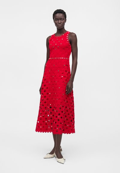 Γυναίκα που φοράει μια κόκκινη μίντι αμάνικη φόρεμα με δαντέλα με τρύπες και λευκές μυτερές γόβες, στέκεται μπροστά σε ένα απλό φωτεινό φόντο.