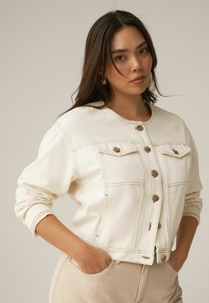 Frau mit langen, dunklen Haaren, die eine cremefarbene, geknöpfte Jacke und beige Hosen trägt, mit den Händen in den Taschen vor einfarbigem Hintergrund stehend.