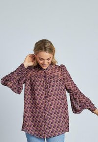 Blusa morada con estampado y mangas abullonadas, que presenta un cuello alto y detalle de botones en la parte delantera. Fabricada en una tela ligera y transparente.