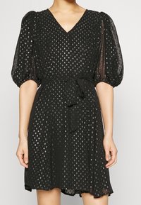 Black dress with a V-neck, short puff sleeves, decorative sequins, belted waist, and a flowy skirt. Fabric is sheer with a textured appearance.