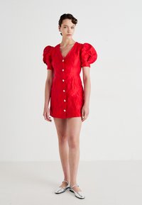 Rød kort kjole med puffede ærmer, knap foran og tekstureret blomstermønster. Modellen bærer sølvfarvede ballerinaer og står foran en ensfarvet baggrund.