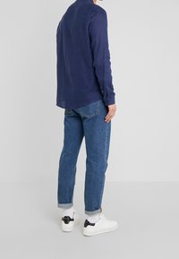Uomo che indossa jeans blu arrotolati, calzini bianchi, sneaker bianche e una maglietta a maniche lunghe blu navy, in piedi contro uno sfondo grigio chiaro semplice.