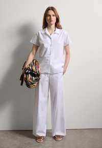 Weißes kurzärmliges Shirt und weit geschnittene Hose aus leichtem Stoff; das Modell hält eine bunte Stricktasche mit einem braunen Tragegurt.