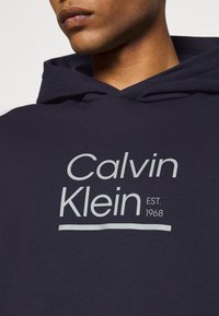 Felpa con cappuccio blu scuro caratterizzata da un grande logo bianco "Calvin Klein" con "EST. 1968" sottostante, realizzata in un morbido tessuto testurizzato.
