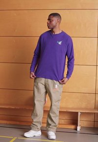 Maglietta Nike viola a maniche lunghe con logo, abbinata a pantaloni beige con bottoni laterali e branding NBA. Sneakers bianche completano il look.