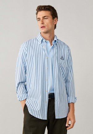 Hombre con camisa de botones a rayas azul claro sobre un polo azul claro, con pantalones oscuros, de pie con una mano en el bolsillo sobre un fondo liso.