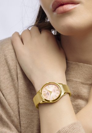 Montre avec un bracelet en maille dorée et un cadran en rose soleil, avec des index horaires dorés et un design de boîtier élégant. Portée au poignet.