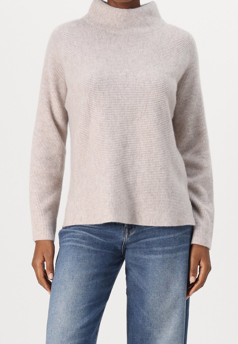 Personne portant un pull à col roulé côtelé beige clair et un jean bleu, debout devant un fond blanc uni.