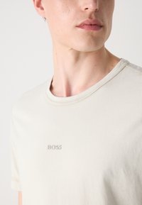 Bežinis medvilnės marškinėliai su apvaliu kaklu ir trumpomis rankovėmis. Yra subtilus iškaltas "BOSS" logotipas ant krūtinės. Minkšta tekstūra, neformalus dizainas.