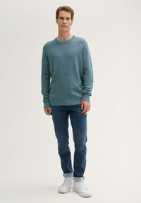 Blauer Strickpullover mit rundem Ausschnitt, kombiniert mit hochgerollten dunkelblauen Jeans und weißen Sneakers. Einfaches Design, glatte Textur.