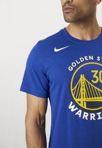 Tricou din bumbac albastru cu un grafic rotund ce prezintă un design cu un pod galben și textul "GOLDEN STATE WARRIORS" pe față, logo-ul Nike pe umăr.