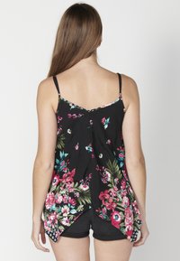 Top nero floreale con spalline sottili, caratterizzato da fiori vivaci in rosa e verde acqua, un design fluente e un orlo asimmetrico. Indossato con pantaloni corti neri.