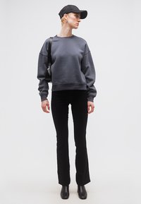 Άτομο που φοράει μαύρο καπέλο, σκούρα γκρι φούτερ, μαύρο στενό παντελόνι, μαύρες μπότες και κρατά μια μαύρη τσάντα ώμου, στέκεται μπροστά σε λευκό φόντο.