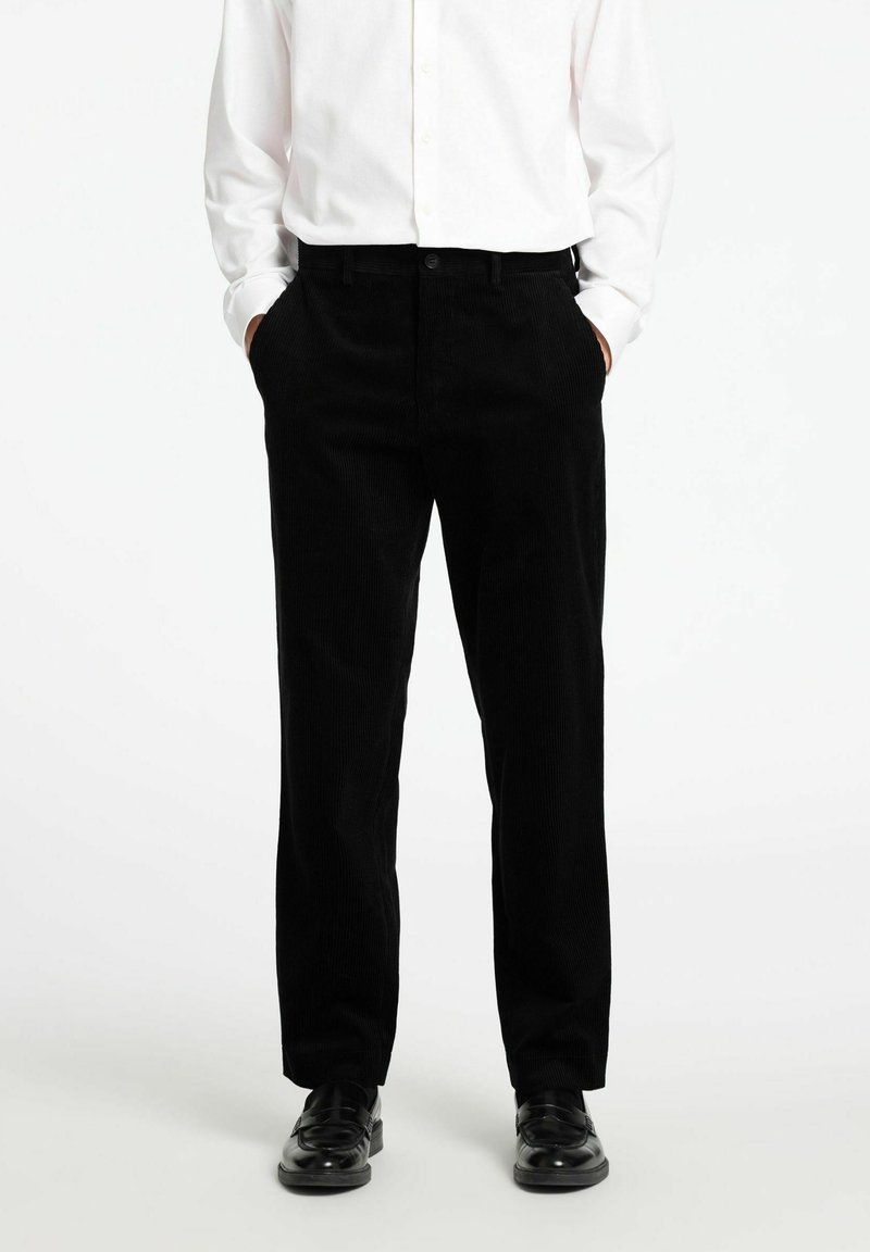Črne kord volna hlače s ravnim krojem, z elastiko v pasu, sprednjimi žepi in subtilno vertikalno teksturo. Usklajene z črnimi čevlji.