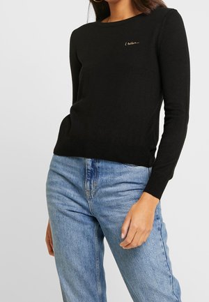 Schwarzer Pullover mit langen Ärmeln und Rundhalsausschnitt, verziert mit dezentem goldenen Sticktext auf der Brust, kombiniert mit hellblauen, hochtaillierten Jeans.