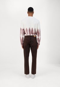 Jersey blanco con patrón de tie-dye en color burdeos, jeans de mezclilla marrones y zapatillas blancas; visto desde la parte trasera contra un fondo liso.