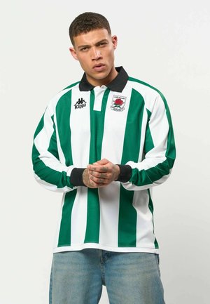 Mand med kort krøllet hår, iført grøn- og hvidstribet rugbytrøje med sort krave og blå jeans, stående mod en ensfarvet baggrund.