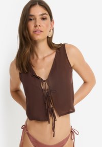 Καφέ αμάνικη μπλούζα με V-λαιμόκοψη και κλείσιμο με κορδόνι, κατασκευασμένη από ελαφρύ ύφασμα. Διαθέτει άνετη εφαρμογή και μίνιμαλ σχεδιασμό.