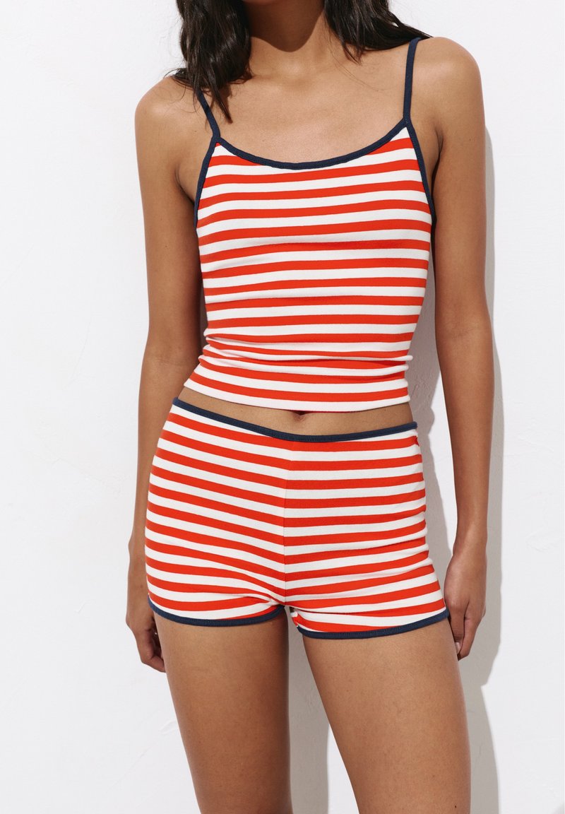 Femme portant un débardeur à rayures horizontales rouges et blanches et un short assorti avec une bordure foncée sur un fond blanc uni.