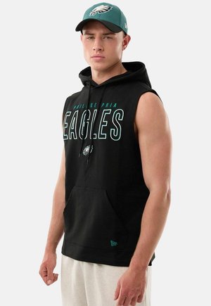 Junger Mann trägt einen ärmellosen schwarzen Philadelphia Eagles Hoodie und eine grüne Eagles Baseballkappe, steht vor einem schlichten Hintergrund.