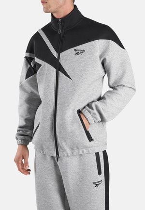 Man draagt een grijs-zwart Reebok trainingspak met een jas met rits en bijpassende broek, handen gedeeltelijk in de zakken, kijkt naar links.