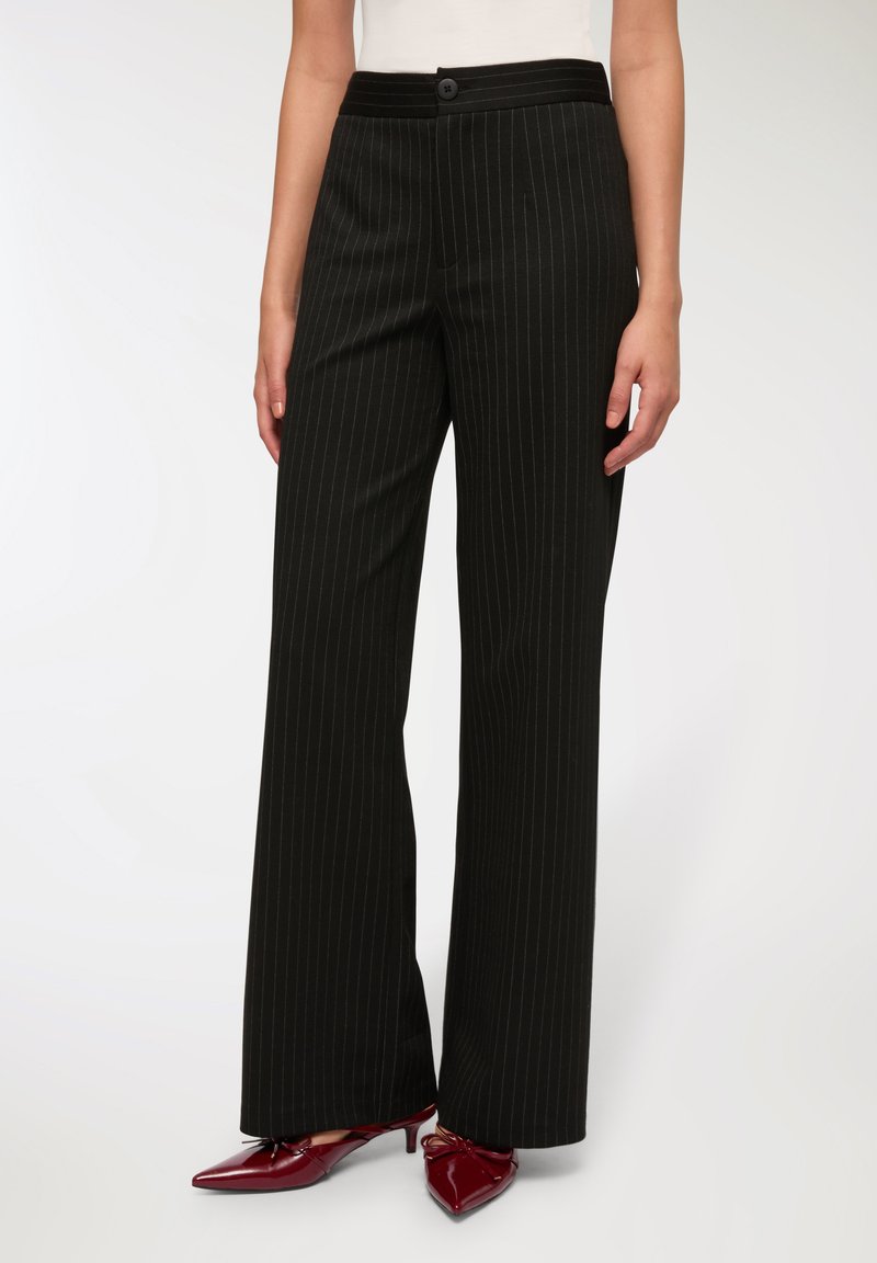 Czarne spodnie w cienkie paski o luźnym kroju, z zapięciem na guzik w talii i gładką fakturą. Połączone z błyszczącymi burgundowymi butami.