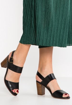 Czarne skórzane sandały na wysokim obcasie z odkrytą palcą, regulowanym paskiem wokół kostki oraz masywnym drewnianym obcasem. Widoczny detal z boku z klamrą.