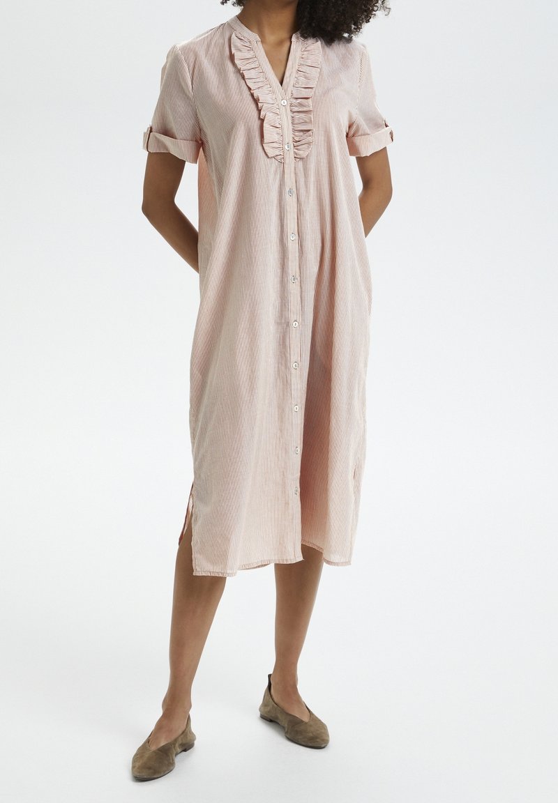 Femme portant une robe rose clair, longueur genoux, boutonnée, avec manches courtes retroussées et un volant au niveau de l'encolure, associée à des ballerines marron.