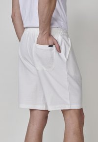 Uomo che indossa pantaloni corti bianchi a trama con vita elasticizzata e tasca posteriore, con una mano appoggiata all'interno della tasca.