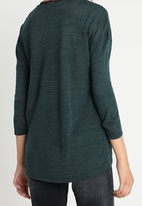 Haut léger vert foncé à manches longues avec une coupe décontractée, un ourlet arrondi et une texture subtile. Vue de dos montrant des manches 3/4 et des coutures minimales.