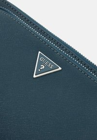 Portafoglio con superficie testurizzata color teal e hardware argentato, caratterizzato da un logo triangolare con "GUESS" e un punto interrogativo, e dettagli in zip argentati.