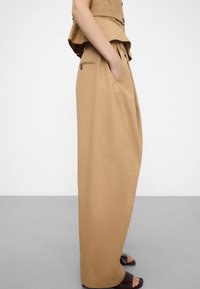 Woman wearing high-waist wide-leg beige trousers with pockets, paired with brown slide sandals, standing against a plain background.