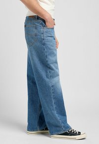 Personne portant un jean en denim bleu ample avec une main dans la poche et des baskets en toile noir et blanc, se tenant contre un fond uni.