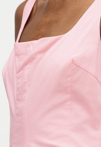 Κοντινό πλάνο ενός ατόμου που φοράει ροζ ανοιχτό ροζ αμάνικη μπλούζα με ευθεία γραμμή λαιμόκοψης και εμφανείς ραφές.